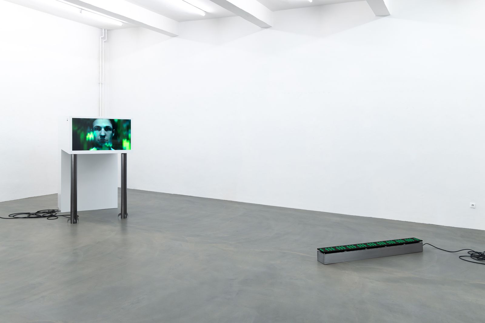 an installation with 2 sculptures in a white cube space, one sculpture has an embedded screen showing a man's face and the other has a matrix screen on top
